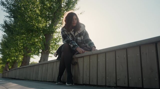 Young woman with long hair wears plaid jacket over black tights while sitting outdoors. Woman holds open notebooks on her lap as sunlight filters through trees creating soft lighting