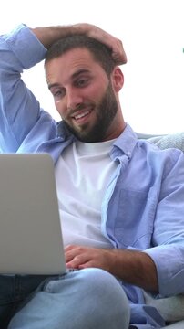 Casual young man with a beard sitting comfortably on the sofa while engaging in a video call on his laptop, smiling and waving, showing a range of positive emotions in a bright living room