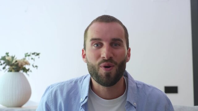 Smiling handsome man with a beard waving hello and enthusiastically talking to the camera, actively using hand gestures during a pleasant video conference call while sitting on the sofa