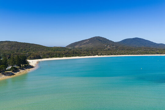 Aerial view of the expansive sandy shoreline and clear turquoise water of Front Beach, with coastal vegetation and mountains rising in the background near South West Rocks, New South Wales, Australia.