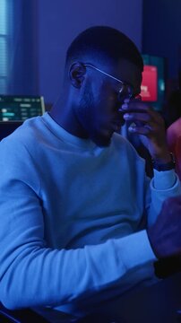 Vertical shot of young adult African American man rubbing tired eyes while programming at computer monitor. Clip suits tech burnout campaigns, eye care ads and cybersecurity training videos