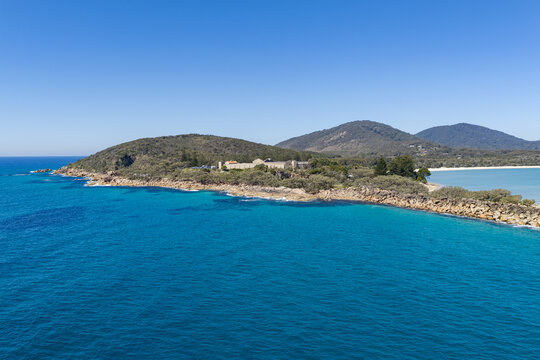 An aerial view of Laggers Point surrounded by clear turquoise ocean, with the historic Trial Bay Gaol visible in the background. 