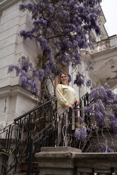 Blooming wisteria frames a young woman posing gracefully on iron stairs outside a historic building