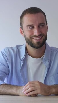 Handsome young man with a short beard sitting at a wooden table, smiling shyly while looking away and then at the camera, showing a hint of bashfulness and embarrassment in his expression