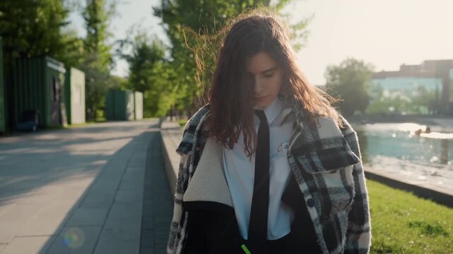 Girl writes notes on paper by hand while seated outside beside water feature. Girl wears casual attire and carries personal items surrounded by greenery and distant buildings under bright