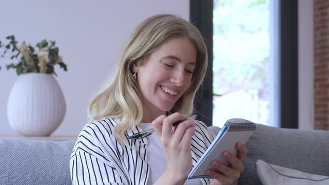 Focused young woman sitting on a comfortable sofa at home, thoughtfully writing down ideas, plans, or a to-do list in a notepad while smiling and brainstorming for inspiration