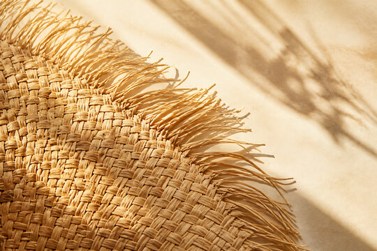 A close-up of a straw or raffia woven hat with long fringe against a sunlit, textured background.