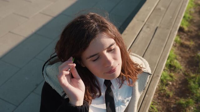 Young adult female stands outdoors on paved area beside wooden planks wearing white collared dress shirt and black necktie. Woman holds various items near her ear including pens or phones