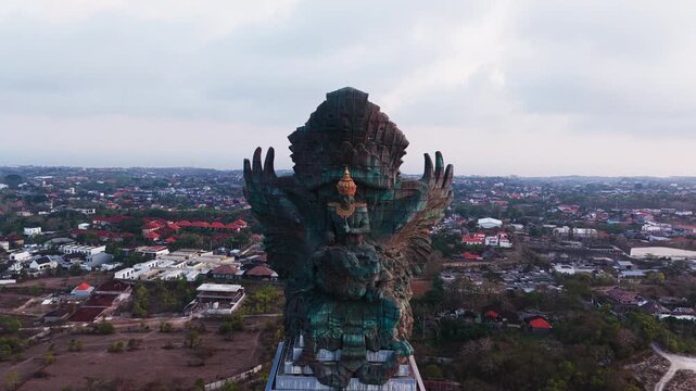 Bali cultural landmark Garuda Wisnu Kencana colossal bronze and copper statue backlit by golden hour light towers over developing urban landscape displaying Hindu sculpture in aerial drone view