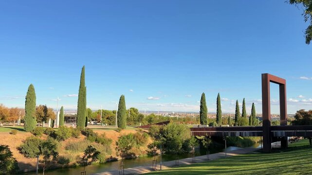 Peaceful panoramic view of Juan Carlos I Park in Madrid, Spain, featuring lush green lawns, a gentle river, and the city skyline under a clear blue sky