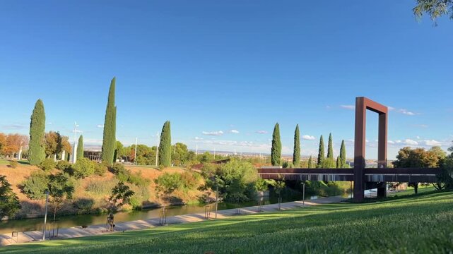 TIMELAPSE | Beautiful sunny day at the Felipe VI Forest Park in Valdebebas, Madrid, with people walking their dogs along the river near a modern corten steel bridge under a clear blue sky