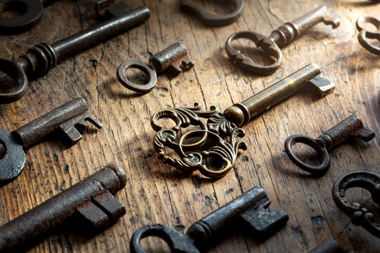 Collection of antique keys lying on a rustic wooden surface, with a dramatic beam of light highlighting the most ornate brass key. Atmospheric vintage still life evoking mystery, history.