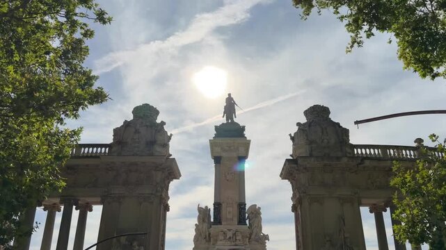 Sunny day illuminating the monument to Alfonso XII in the Retiro Park, Madrid, Spain, a famous tourist landmark framed by green trees against a bright sky with some clouds