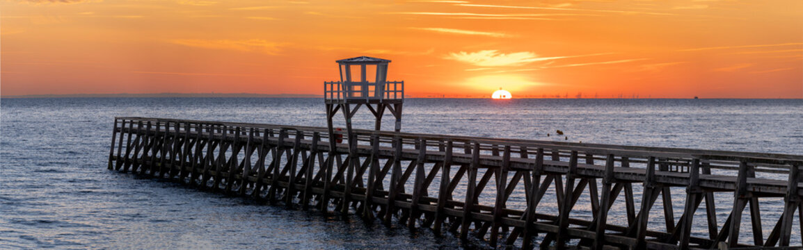 Luc-Sur-Mer, France - 08 08 2026:  Panoramic view of a wooden pier in front of the beach, sea, a colorful sunrise and Le Havre city far away