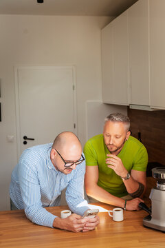 Gay couple drinking coffee and looking at a smartphone together in the home kitchen