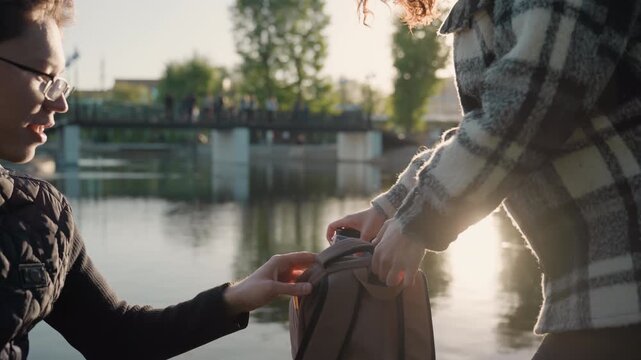 Two people pass notebook objects between hands beside calm waterway with soft sunlight. One person receives and places items into another backpack suggesting organized handoff of study