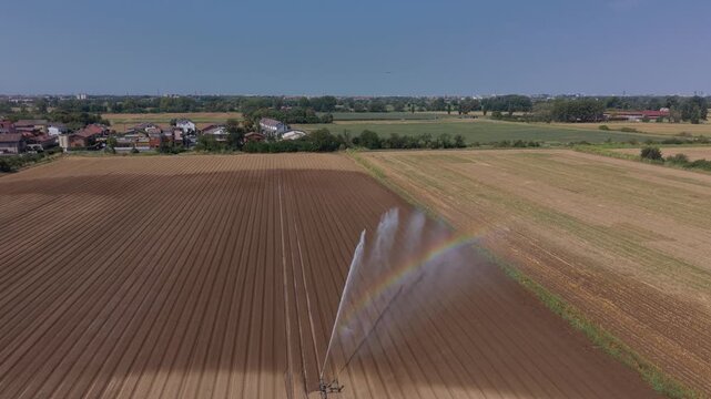 Aerial drone view of a large gun sprinkler watering a newly planted vegetable field in Lombardy, Italy, with a subtle rainbow forming in the spray. Suitable for agriculture and irrigation content.
