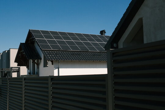Residential house with large rooftop solar panel array behind a modern fence under clear blue sky, representing renewable electricity, energy independence and sustainable living.