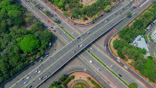 Aerial view of a multi-level highway interchange with cars driving on asphalt roads surrounded by lush green trees and urban vegetation in Jakarta, Indonesia.