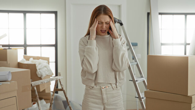 Woman holding temples and grimacing amid stacked moving boxes, packing paper and ladder in a sunlit room inside a building; moving stress.