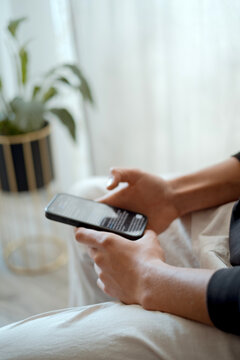 Hands hold phone, fingers tapping gently. Soft light filters through sheer curtains behind. Potted plant adds touch of green to quiet room. Person sits comfortably, focused on digital device