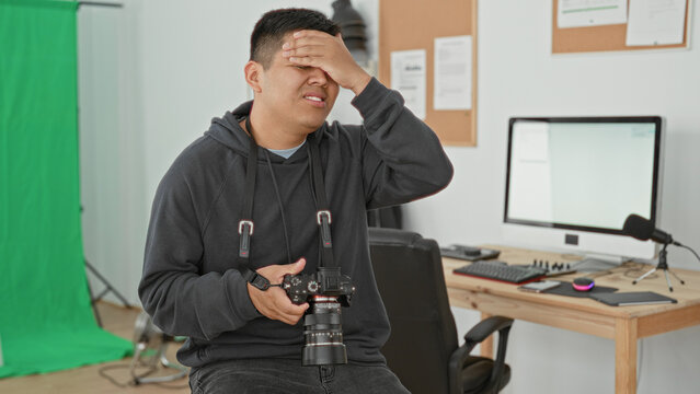 Man photographer holding camera and checking controls with hand on forehead beside green screen and computer desk in studio; frustration problem solving.