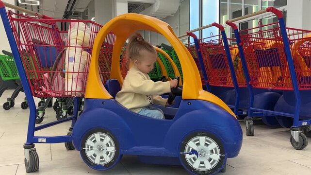 A girl pretends to drive a blue and yellow shopping cart car inside a store, playful shopping, childhood imagination, retail convenience.