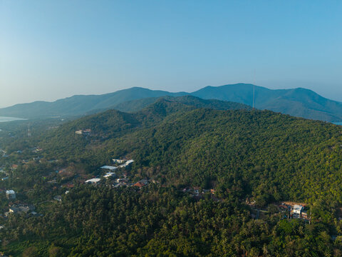 Aerial view of Karimunjawa hills covered in dense tropical forest with a small village nestled at the base under a clear blue sky in Karimunjawa, Central Java, Indonesia.