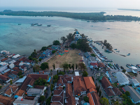 Aerial view of Karimunjawa Town Square, featuring traditional terracotta-roofed houses, a busy harbor with small boats, and turquoise waters in Karimunjawa, Central Java, Indonesia.