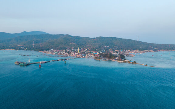 Aerial view of Karimunjawa town square and the long pier extending into the turquoise sea, surrounded by lush green hills and clear blue water in Karimunjawa, Central Java, Indonesia.