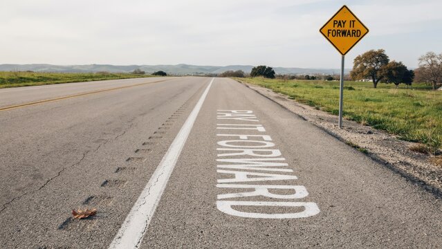 An empty country road features 'Pay It Forward' painted on the asphalt and a matching yellow sign. Ideal for kindness campaign concepts.