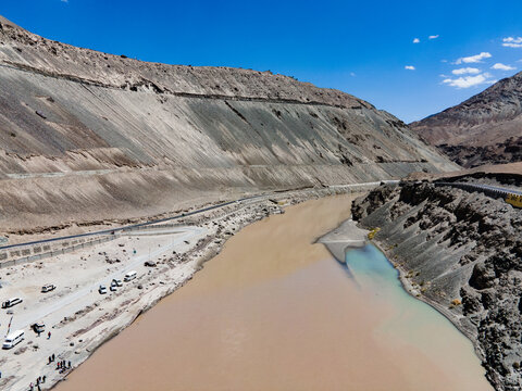 Aerial view of the confluence of the Indus and Zanskar rivers, showing the meeting of muddy and turquoise waters amidst rugged mountains and a road in Ladakh Division, Ladakh, India.