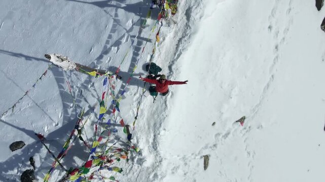 Aerial drone footage rising above a hiker with outstretched arms among colorful prayer flags on snowy Tserko Ri summit, Langtang National Park, Nepal Himalayas, near 5000m altitude