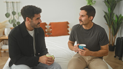 Two men sit on a bed holding coffee mugs, one gestures with his cup while facing the other in a...