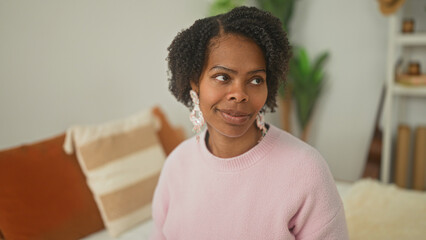 Fototapeta premium Young woman on bed wearing pink sweater and statement earrings, eyes closed with pursed lips beside pillows and plant in building; calm mindfulness.