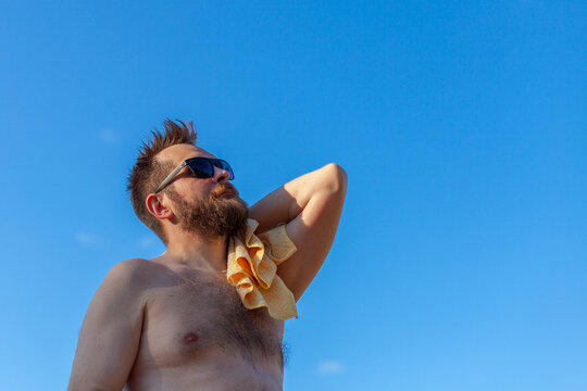Shirtless bearded man wearing sunglasses holding yellow towel against clear blue sky. Summer leisure and relaxation. Enjoying summer, vacation, lifestyle, sunbathing, travel.