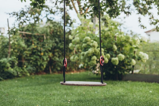 Empty wooden swing hanging by black ropes in  green backyard. Сhildhood memories, summer moments, relaxation, outdoor living, rustic charm and family time.