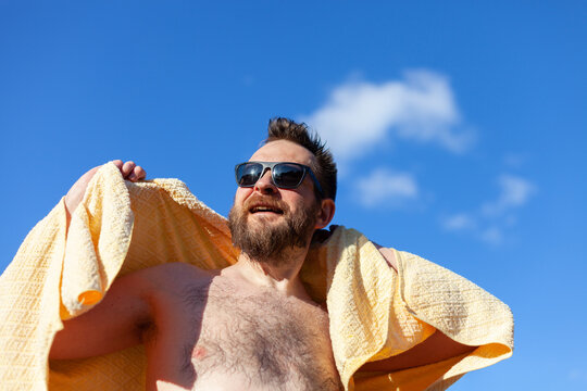 Happy bearded man with sunglasses holding towel over shoulders against bright blue sky. Summer vacation, relaxation, positive emotions, travel, wellness, and beach leisure. Man enjoying sunny weather.
