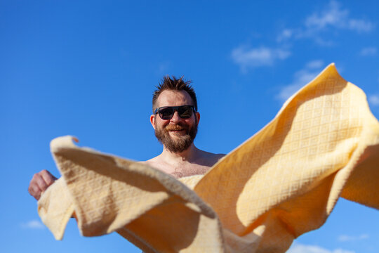 Happy bearded man wearing sunglasses holding large yellow towel in motion against clear blue sky. Man enjoying sunny day at beach or pool. Summer relaxation, vacation vibes, positive emotions.