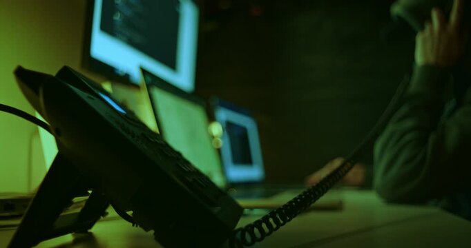 Defocused person in a dark hoodie making a telephone call while sitting in a dim room surrounded by glowing screens and laptops. Telephone fraud, scams, cybercrime, deception, identity theft, hacking