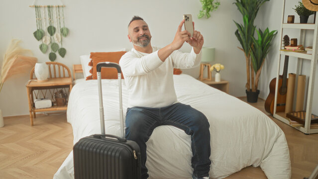 Man seated on bed with carryon suitcase, holding smartphone with both hands taking a selfie in a bedroom inside a building; anticipation travel.