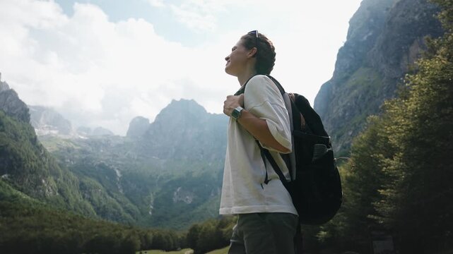 Young female traveler with a backpack enjoying a breathtaking mountain view while standing in a scenic meadow, capturing freedom, adventure and connection with nature.