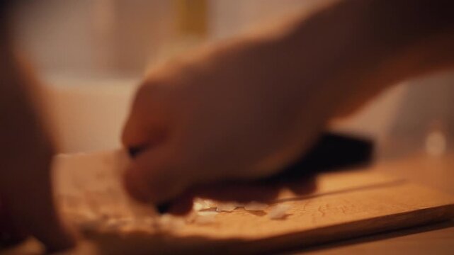 In dim kitchen, chef chops onions on a wooden cutting board, evening shadows casting a culinary mood as the cook prepares a flavorful dish. The knife glints under faint light, showing precise slices