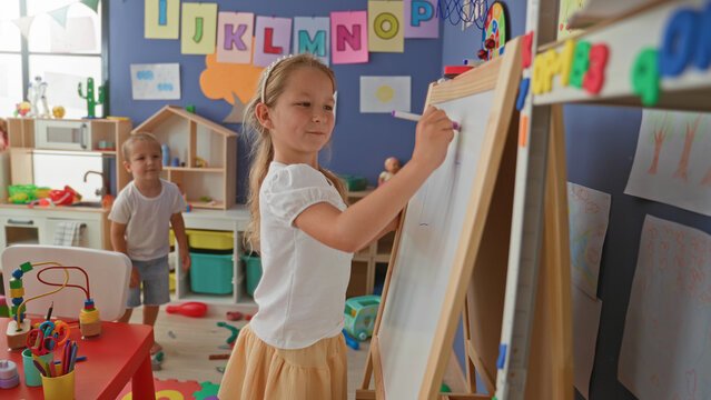 Girl writing on easel whiteboard with marker while brother watches in a preschool playroom building; playful sibling learning.