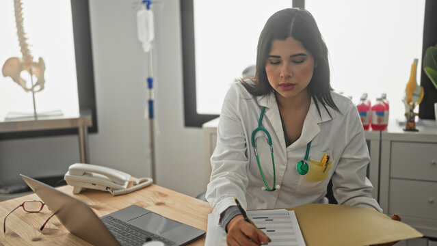 Woman doctor with green stethoscope writing notes on a patient chart at desk with laptop and phone in building; focused medical care.