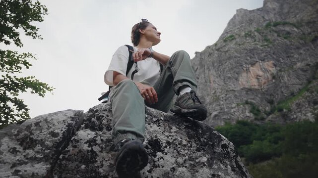 Young female traveler with a backpack sitting on a large rock and enjoying a breathtaking mountain view, capturing freedom, adventure, relaxation and connection with nature.