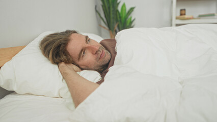 Man lying in bed with hand behind head on pillow under white duvet cover, bedroom  serenity contemplation. © Krakenimages.com