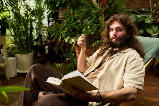 Portrait of young adult Caucasian man holding mug and reading book among indoor house plants. Suitable for mental wellbeing, relaxation and mindfulness lifestyle campaigns