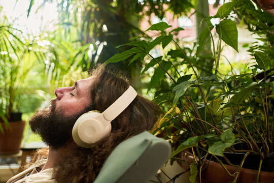 Caucasian young adult man resting with closed eyes, listening to audio through wireless headphones near house plants. Image suitable for mental wellbeing and relaxation apps promotion