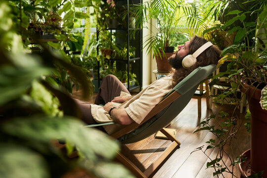 Young adult Caucasian man resting in chair wearing headphones among indoor plants, relaxing during audio meditation. Image useful for mental wellbeing and relaxation campaigns